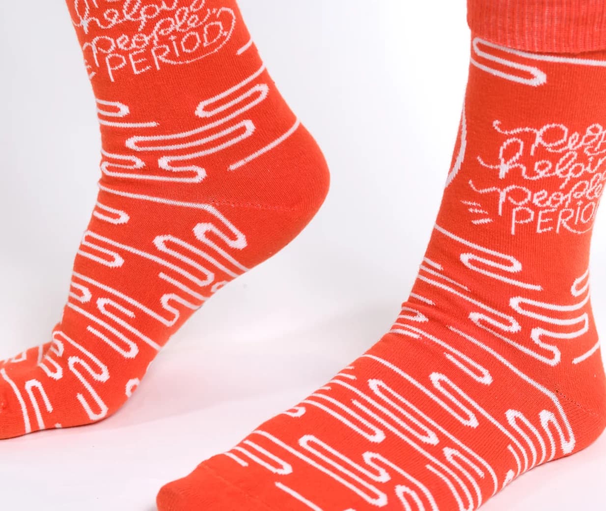 A pair of bright orange crew socks with white wavy line patterns and the text 'Help! Help! Perfect Period.' knitted onto the side, shown on feet against a clean white background.
